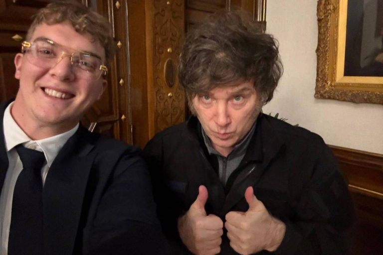 A imagem mostra duas pessoas tirando uma selfie. A pessoa à esquerda tem cabelo curto e usa óculos com armação dourada, vestindo um terno escuro e gravata. A pessoa à direita tem cabelo bagunçado e está fazendo um gesto de