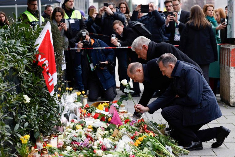 A imagem mostra um grupo de pessoas em um local de homenagem, onde há uma grande quantidade de flores e velas dispostas no chão. Duas pessoas estão se agachando para colocar flores, enquanto outras pessoas ao fundo estão tirando fotos com seus celulares. Algumas pessoas estão vestindo coletes de segurança. O ambiente parece ser urbano, com um cercado ao fundo.