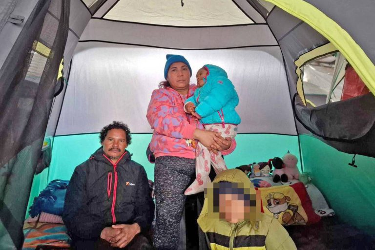 A imagem mostra uma família dentro de uma barraca. Um homem está sentado no chão à esquerda, enquanto uma mulher em pé segura uma criança pequena. A mulher está vestindo um casaco rosa e a criança está vestida com um casaco azul. À direita, uma outra criança está parcialmente visível, usando um casaco amarelo. O interior da barraca é iluminado e apresenta um fundo verde.