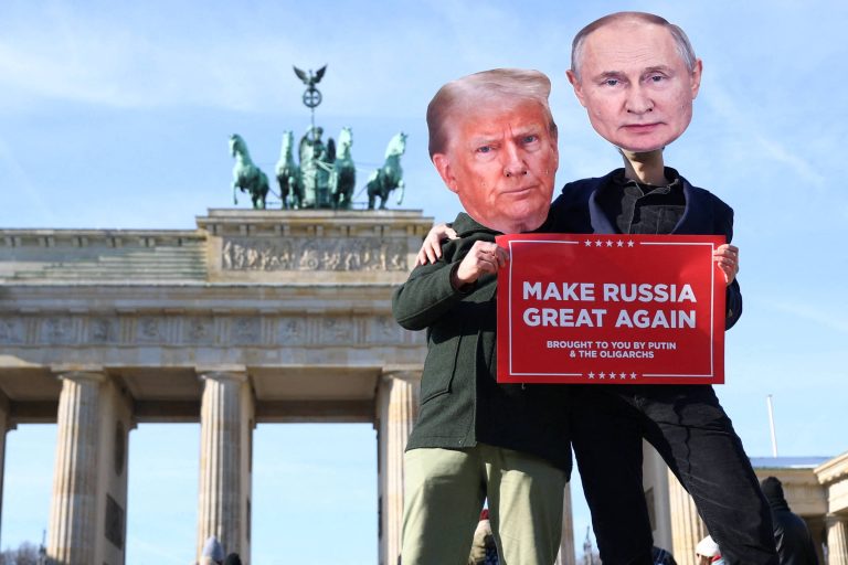A imagem mostra duas pessoas em frente ao Portão de Brandemburgo, em Berlim. Uma delas está vestindo uma máscara com o rosto de Donald Trump e a outra com o rosto de Vladimir Putin. Ambas seguram um cartaz vermelho que diz