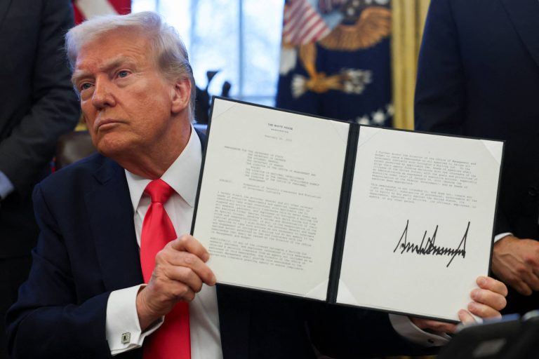 A imagem mostra Trump, um homem branco com cabelo loiro e uma gravata vermelha, sentado em uma mesa. Ele está segurando um documento em uma pasta, exibindo sua assinatura. Ao fundo, há uma janela com cortinas e bandeiras dos Estados Unidos.