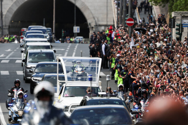A imagem mostra uma grande multidão ao longo de uma rua, com pessoas acenando e tirando fotos. À frente, há uma carreata de veículos, incluindo um carro com um teto transparente, que parece estar transportando uma figura importante. Policiais em motocicletas estão na frente da carreata, e há um túnel ao fundo. A atmosfera é de celebração e entusiasmo.