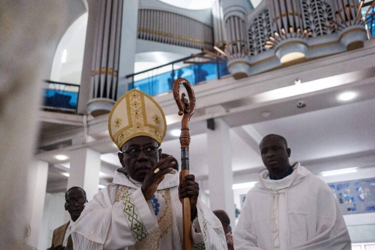 A imagem mostra um homem vestido com vestes litúrgicas, usando um grande chapéu amarelo e segurando um cajado. Ele está em uma igreja, com um órgão visível ao fundo. Ao lado dele, há outro homem vestido de branco. O ambiente é iluminado e possui uma arquitetura moderna.