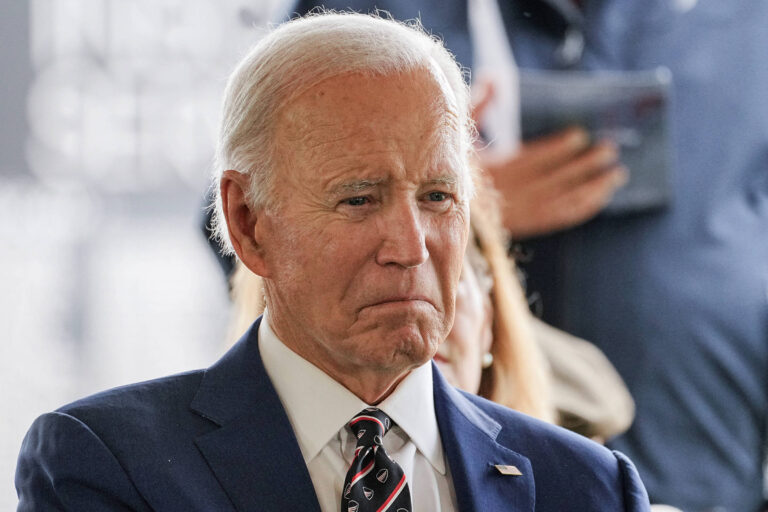 Joe Biden é um homem idoso e com cabelo branco. Ele está uma expressão séria e usando um terno escuro e uma gravata. Ao fundo, há pessoas e um ambiente que parece ser um evento público.