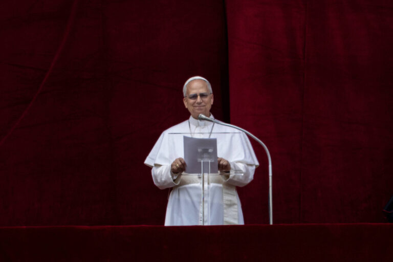 Um homem vestido com vestes papais brancas, incluindo uma batina e um manto, está em pé em um púlpito. Ele segura um papel e parece estar prestes a fazer um discurso. O fundo é de um tecido vermelho, criando um contraste com suas vestes. O ambiente é formal e solene.