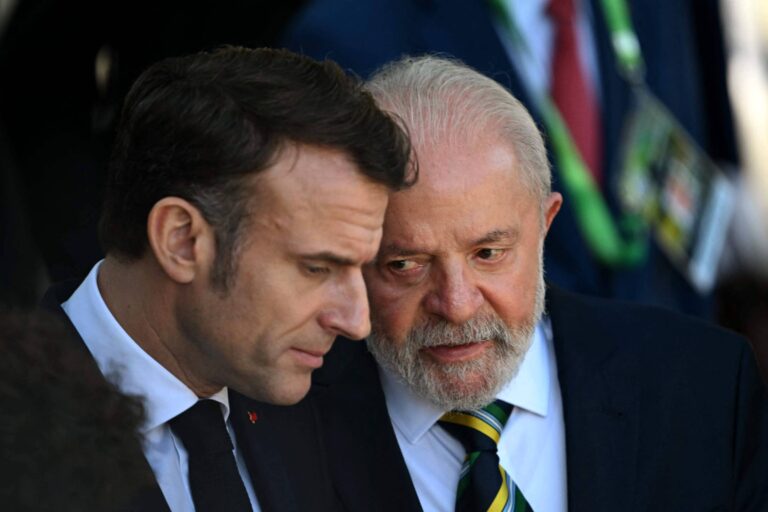 A imagem mostra dois homens em um momento de conversa próxima. O homem à esquerda tem cabelo escuro e usa um terno preto com uma gravata. O homem à direita tem cabelo grisalho e uma barba, vestindo um terno escuro com uma camisa branca e uma gravata colorida. Eles parecem estar em um ambiente formal, possivelmente em um evento político.
