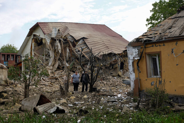 A imagem mostra dois indivíduos caminhando em direção a um edifício parcialmente destruído, com a estrutura do telhado colapsada e as paredes danificadas. Ao fundo, há outro edifício que também apresenta danos significativos. O chão está coberto de escombros e detritos, e algumas árvores e vegetação estão visíveis ao redor.