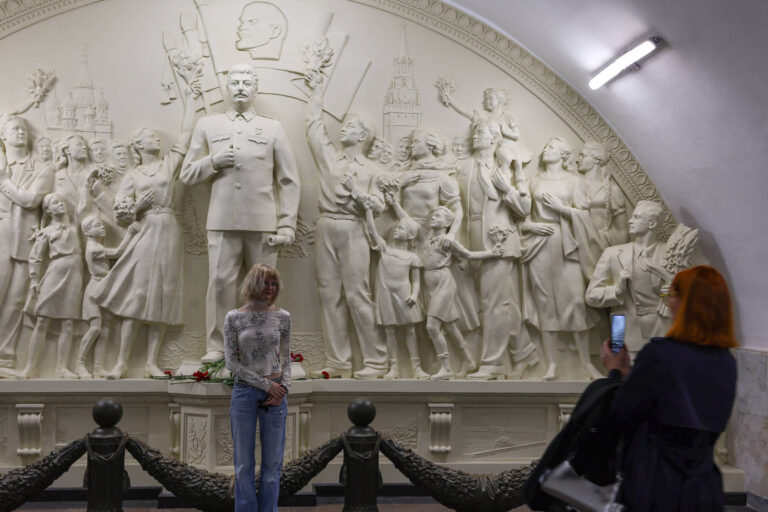 A imagem mostra um monumento em relevo que retrata várias figuras, incluindo um homem em uniforme militar, Josef Stálin, e várias crianças e adultos em poses diversas. Uma mulher está em primeiro plano, de costas, posando para uma foto, enquanto outra mulher, com cabelo ruivo, está tirando a foto com um celular. O ambiente é iluminado artificialmente e as paredes são de um tom claro.