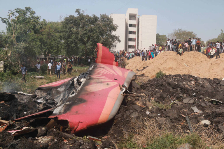 A imagem mostra os destroços de um avião caído em uma área com vegetação. O avião está parcialmente queimado, com a fuselagem visível e uma parte da asa. Ao fundo, há um grupo de pessoas observando a cena, e um edifício branco se destaca. Há também uma pilha de terra ou areia próxima aos destroços.