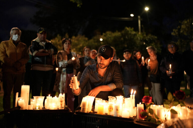 A imagem mostra um grupo de pessoas reunidas em uma vigília noturna, segurando velas acesas. No primeiro plano, um homem está agachado, acendendo uma vela em meio a várias outras velas e rosas dispostas em caixas. Ao fundo, há uma multidão de pessoas observando, algumas com expressões sérias, em um ambiente iluminado por luzes suaves.