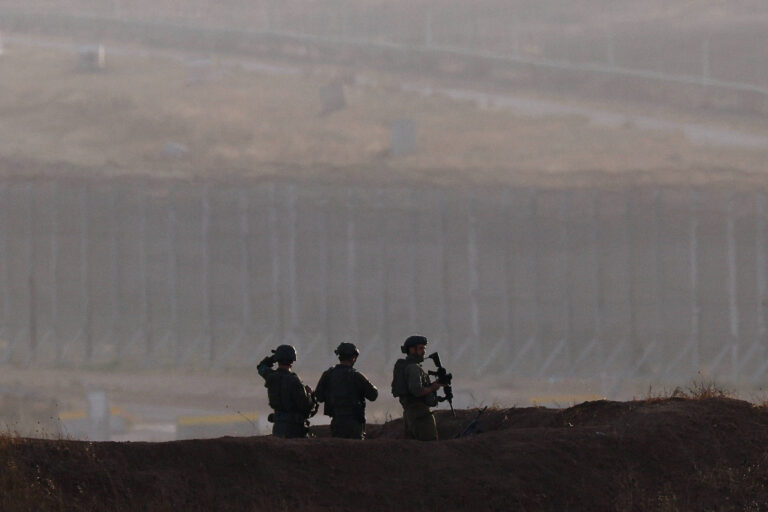 Três soldados estão posicionados em uma área elevada, observando o horizonte. Eles estão vestidos com uniformes militares e portando armas. Ao fundo, há uma cerca alta e uma paisagem nebulosa, sugerindo um ambiente de segurança ou patrulha. A atmosfera é de vigilância e seriedade.