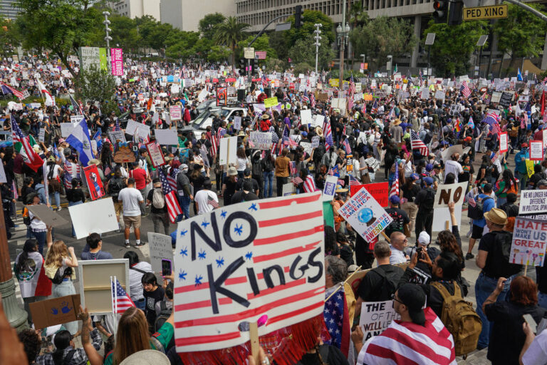 A imagem mostra uma grande multidão reunida em um protesto. Muitas pessoas seguram cartazes e bandeiras, incluindo uma faixa que diz