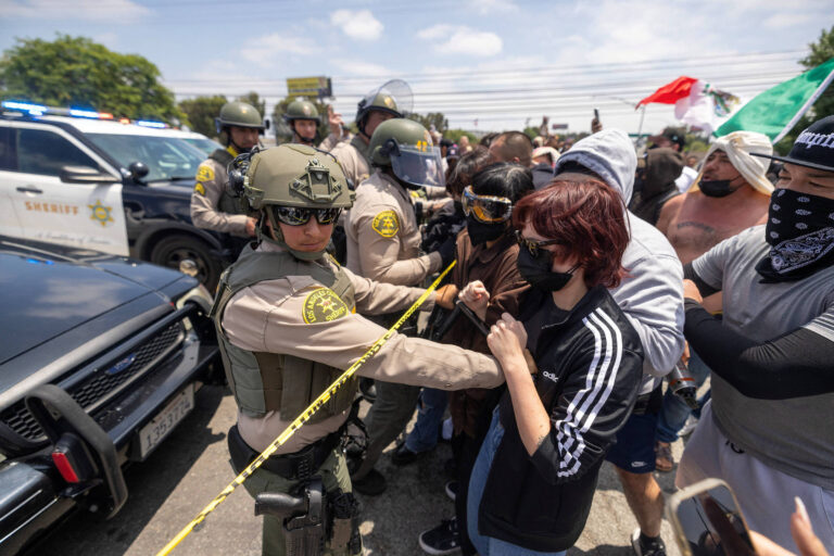 A imagem mostra um confronto entre manifestantes e policiais em um protesto. Policiais em uniformes táticos e capacetes estão posicionados à esquerda, enquanto um grupo de manifestantes, alguns usando máscaras, está à direita. Uma policial está interagindo com uma manifestante, segurando uma fita de isolamento. Veículos da polícia estão visíveis ao fundo, e bandeiras podem ser vistas entre os manifestantes.