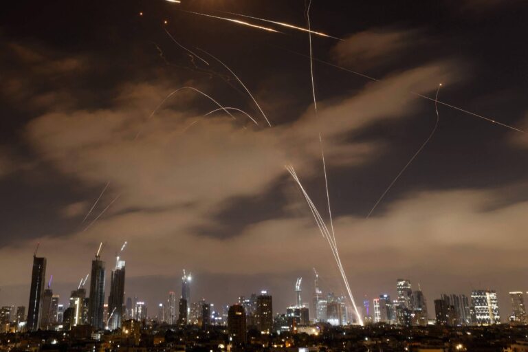A imagem mostra um horizonte urbano à noite, com vários edifícios iluminados. No céu, há várias trajetórias de luz, indicando a presença de mísseis ou foguetes sendo disparados. As nuvens estão visíveis, e a cena transmite uma sensação de atividade intensa no ambiente urbano.