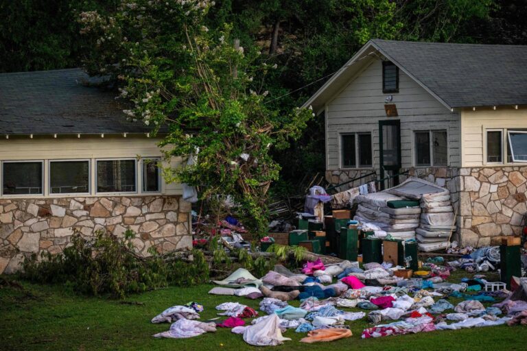 A imagem mostra uma área residencial afetada por um desastre. Há uma casa de dois andares com paredes de pedra e janelas grandes. Na frente da casa, há uma grande quantidade de colchões, roupas e outros itens espalhados pelo chão. Uma árvore está visivelmente danificada, e o ambiente parece desordenado, sugerindo que houve uma situação de emergência ou desastre natural.