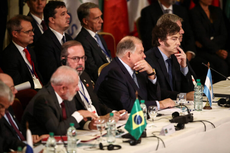 A imagem mostra uma mesa de conferência com vários líderes e representantes sentados. Há bandeiras do Brasil e da Argentina visíveis na mesa. Os participantes estão em trajes formais, alguns usando fones de ouvido. Ao fundo, é possível ver outros homens em pé, com bandeiras de diferentes países ao fundo. A atmosfera parece ser de uma reunião oficial ou cúpula.